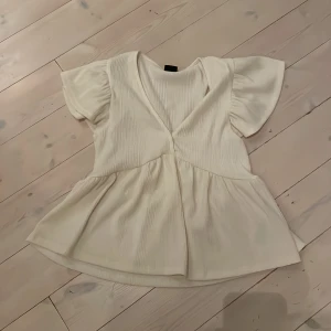 Vit blus med volang från Gina Tricot - Supersöt vit blus från Gina Tricot i storlek XS. Blusen har korta, voluminösa ärmar och v-ringad hals. Materialet är ribbat och ger en snygg struktur. Den har en volang nedtill som ger en flowy och chill vibe. Perfekt för dig som gillar en feminin och trendig stil.