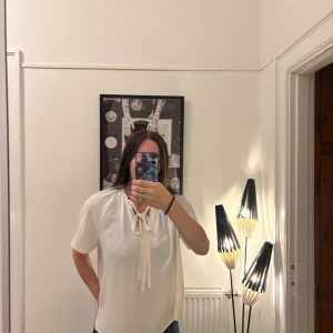 Vit blus med knyt från H&M - Säljer en vit blus från H&M med korta ärmar och snygg knytning vid halsen. Blusen har en lös passform och är tillverkad i ett lätt, flowy polyestertyg som ger en skön känsla. Perfekt att styla med jeans eller kjol för en fräsch look.