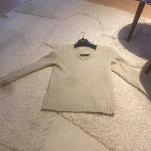 Beige stickad tröja från Vila - Supermjuk beige stickad tröja från Vila med v-ringning och långa ärmar. Perfekt för lager-på-lager och chill dagar. Ribbstickade muddar vid ärmslut och nederkant ger en snygg touch. Enkel att matcha med jeans eller kjol.