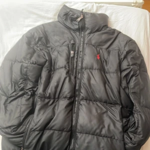 Svart pufferjacka Polo Ralph Lauren M - Svart pufferjacka från Polo Ralph Lauren i storlek M. Jackan har hög krage, dragkedja framtill och en bröstficka med dragkedja. Klassisk röd logga broderad på bröstet och två sidofickor med dragkedja. Perfekt för kyliga dagar.