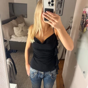 Svart linnetopp med volangdetalj - Säljer en svart linnetopp med smala axelband och snygg draperad urringning. Toppen har volangdetaljer längs sidan som ger en extra touch.