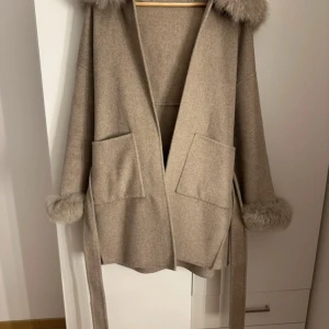 Beige kappa med päls  och bälte - Snygg beige kappa med fluffig päls på huva och ärmslut. Jackan har två stora fickor framtill och ett matchande bälte i midjan för extra stilpoäng. Perfekt för dig som gillar en chill och lyxig vibe under höst och vinter. Jag har bara använt den under en höst termin köpte för 2499 
