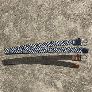 Två flätade bälten  - Säljer två snygga flätade bälten, ett i blått, vitt och grått med mörkblå detaljer och ett i mörkgrått med bruna detaljer. Båda har silverfärgade metallspännen och är gjorda i syntetmaterial med skinnimitation vid spännet. Perfekt för att ge outfiten lite extra stil. Gråa bälte skick 8/10 det blåa bältet skick 10/10. Säljs för 119kr pris kan diskuteras vid snabb affär!