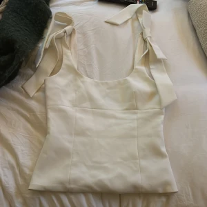 Vit linnetopp med knytband - Säljer en stilren vit linnetopp med breda axelband som knyts i rosetter på axlarna. Toppen har en fyrkantig urringning och är figursydd för en snygg passform. Perfekt att styla med jeans eller kjol för en fräsch look. använd 1 gång 