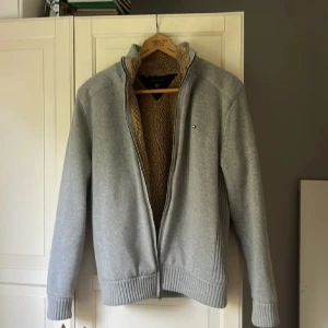Grå fleecekofta Tommy Hilfiger - Mysig grå kofta från Tommy Hilfiger med beige fleecefoder och ribbade muddar. Tröjan har dragkedja framtill, ståkrage och diskret logga på bröstet. Perfekt för kyliga dagar när du vill vara både varm och snygg.