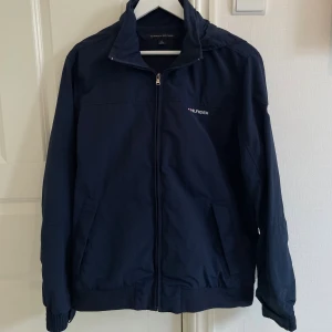 Tommy Hilfiger vindjacka - Snygg mörkblå vindjacka från Tommy Hilfiger med dragkedja framtill, broderad logga på bröstet och tryckt Hilfiger-text på kragen. Jackan har långa ärmar, två fickor och elastiska muddar. Perfekt för vår och höst! Passar dig som är ca 175-187 cm lång. 