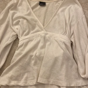 Vit omlottopp med knyt från Gina Tricot - Säljer en vit omlottopp från Gina Tricot i storlek S. Toppen har lång ärm, v-ringad hals och ett snyggt knyt i midjan. Materialet är mjukt och har en lätt ribbad struktur som ger en chill vibe. Perfekt för dig som gillar enkel men stilren look.