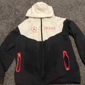 Svart och vit vindjacka Hugo Boss x Mercedes - Snygg vindjacka med huva från Hugo Boss x Mercedes-Benz. Jackan är svart och vit med röda loggor och detaljer, samt röda dragkedjor på fickorna. Materialet är troligtvis polyester och jackan har hel dragkedja framtill. Perfekt för dig som gillar sportig och clean stil.