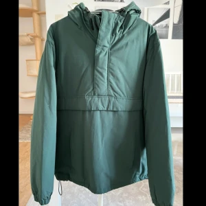 Grön vindjacka från Etirel med huva - Snygg grön vindjacka från Etirel i storlek S, med justerbar huva och dragkedja framtill. Jackan har elastiska muddar vid ärmslut och snörning nedtill för extra passform. Perfekt för dig som gillar enkel och clean stil.Helt ny utan prislapp.