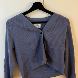 Blå croppad kofta från Zara - Snygg blå croppad kofta från Zara med ribbad struktur och v-ringad hals. Koftan har en knapp framtill och långa ärmar. Perfekt för dig som gillar stilrena och trendiga plagg med lite edge.