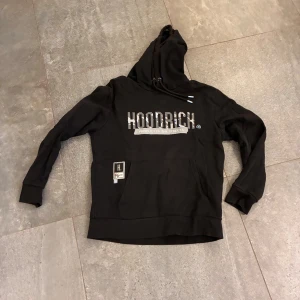 Svart hoodie från Hoodrich - Svart hoodie från Hoodrich med stor logga och text 'Product of the Streets' både fram och bak. Tröjan har huva med snörning, magficka och ribbade muddar. Perfekt för dig som gillar streetwear och vill ha en cool look.                1000kr nypris ungefär från london.