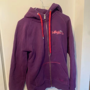 Lila hoodie från Peak Performance - Lila hoodie från Peak Performance med dragkedja, röda snören och broderad logga på bröstet. Mjuk insida och känguruficka framtill. Perfekt för chill dagar eller när du vill ha en färgklick i garderoben.