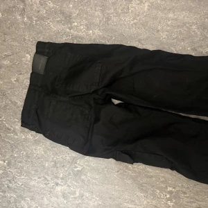 Svarta cargopants med fickor - Svarta cargopants med flera praktiska fickor på sidorna och bak. Byxorna har en avslappnad passform och är tillverkade i ett slitstarkt bomullsmaterial. Perfekta för dig som gillar streetwear och vill ha en cool och funktionell look. Har ett par i storlek S och ett par i M
