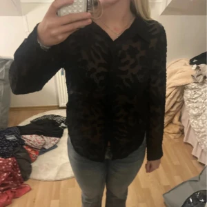 Svart skjorta med leopardmönster - Säljer en transparent svart skjorta med coolt mönster. Skjortan har krage och långa ärmar, perfekt för dig som vill sticka ut lite extra. Snygg att styla med jeans eller kjol för en trendig look.