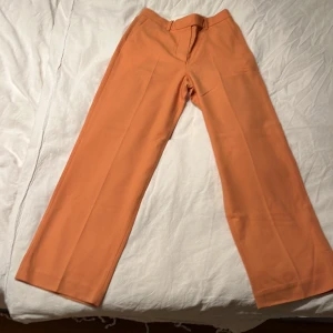 Persikofärgade kostymbyxor strl 34 - Snygga persikofärgade kostymbyxor i rak modell med pressveck. Byxorna har hög midja och stängs med dragkedja och hake. 