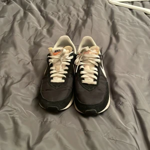 Svarta Nike sneakers strl 42,5 - Säljer ett par klassiska svarta Nike sneakers med vit swoosh och vita snören. Skorna har en retro vibe med svart ovandel i syntet och mockadetaljer, samt en vit mellansula och svart yttersula med rutmönster. Perfekta för dig som gillar stilrena och tidlösa sneakers.