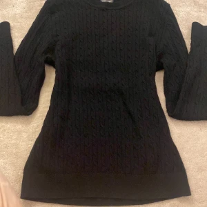 Svart stickad tröja från Crocker - Svart kabelstickad tröja från Crocker i storlek S. Tröjan har rund hals, långärmade ribbade muddar och ett klassiskt flätmönster över hela plagget. Perfekt för dig som gillar enkel och stilren look med lite struktur.