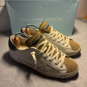 Philippe model skor - Snygga Philippe Model sneakers i vitt skinn och beige mocka med mörkblå detalj på hälen. Klassisk låg modell med snörning och rund tå. Sulan har tydlig 'MODEL'-logga undertill. Tillverkade i Italien och perfekta för dig som gillar stilrena men unika sneakers.