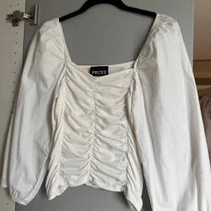 Vit blus med rynk från Pieces - Säljer en vit blus från Pieces med snygg rynkdetalj framtill och puffiga långärmar. Blusen har fyrkantig halsringning och är croppad i modellen. Perfekt att styla med höga jeans för en trendig look.