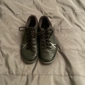 Svarta Nike fotbollsskor strl 38,5 - Säljer ett par svarta Nike fotbollsskor med silverdetaljer och snörning. Skorna har en låg profil och är tillverkade i syntetmaterial med mönstrad sula för bra grepp på planen. Perfekta för dig som vill ha stilrena och funktionella skor till inomhusfotboll.