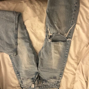 Bootcut ljusblå jeans med slitning - Ljusblå bootcut jeans med låg midja och rejäl slitning på ena benet. Jeansen har klassisk femficksdesign och är tillverkade i denim. Perfekta för dig som gillar en avslappnad och edgy stil.