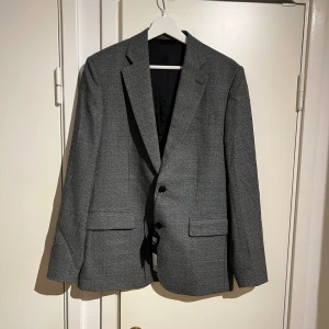 Grå kavaj från J.Lindeberg  - Säljer en skit snygg och stilig grå kavaj från J.Lindeberg i klassisk modell med två knappar framtill och fickor med lock. Även innefickor. Kavajen har ett diskret mönster och är fodrad med svart tyg. Perfekt för dig som vill ha en stilren look med modern touch perfekt för vardagen men även när man skall vara uppklädd, Nypris på kavajen ligger runt 2500kr-3000kr och är storlek 46r vilket motsvarar en medium. Bredd över axlarna är 45cm och längden är 70cm. Passar dig som är mellan cm 175-182cm