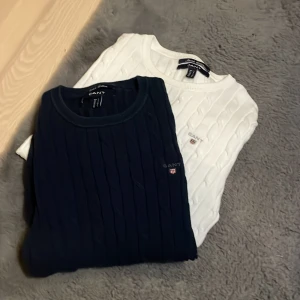 Marinblå och vit kabelstickad GANT-tröja - Två snygga kabelstickade tröjor från GANT i stretchig bomull, en marinblå och en vit. Båda har rund hals, lång ärm och broderad GANT-logga med sköld på bröstet. Perfekt för dig som gillar klassisk och clean stil.