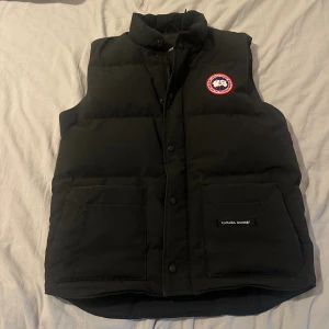 Äkta! Svart Canada Goose freestyle väst - Svart dunväst från Canada Goose med klassisk logga på bröstet. Västen har hög krage, två stora fickor framtill och stängs med både dragkedja och knappar. Perfekt för lager-på-lager och riktigt snygg till streetwear. Original pris ca 8000kr