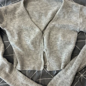 Grå croppad kofta med knappar - Supermysig grå kofta med croppad passform och djup v-ringning. Tröjan har ribbade muddar och stängs med tre knappar framtill. Perfekt att slänga över en topp för en chill look.