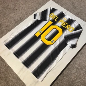 Juventus Del Piero #10 Nike tröja M - Juventus matchtröja med Del Piero och nummer 10 på ryggen. Svart och vit randig design, gult tryck, Juventus klubbmärke med två stjärnor, Betclic sponsor på bröstet och Nike swoosh. Tillverkad i lätt, ventilerande polyester med Dri-Fit teknologi.