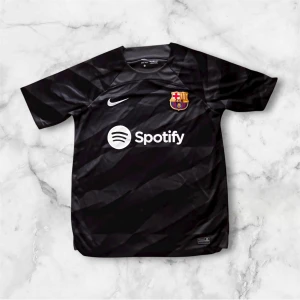 Svart FC Barcelona fotbollströja Nike - Snygg svart FC Barcelona fotbollströja från Nike med klubbmärke på bröstet och Spotify-logga framtill. Tröjan är tillverkad i 100% polyester med Dri-Fit-teknologi och har korta ärmar. Diskret randigt mönster och UNHCR-logga på ryggen. Perfekt för dig som älskar Barca! Nypris ca 700kr: Mitt pris 299kr!