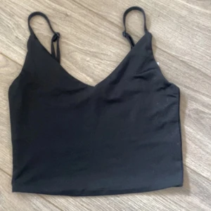 Svart croppad linnetopp - Snygg svart croppad linnetopp med smala justerbara axelband och v-ringad hals. Perfekt att styla med jeans eller kjol för en trendig look. Materialet är mjukt och följsamt, vilket gör toppen bekväm att bära hela dagen.