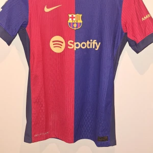 FC Barcelona Lamine Yamal matchtröja - Säljer en officiell FC Barcelona fotbollströja från Nike med Lamine Yamal och nummer 19 på ryggen. Tröjan är kortärmad, har klassisk röd och blå färgdelning, gult tryck och Dri-Fit ADV-material. Sponsorer som Spotify och UNHCR samt Champions League-märke på ärmen.