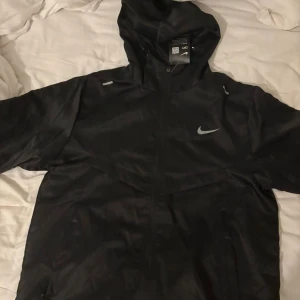 Svart Nike vindjacka med huva XL - Nike vindjacka i svart med huva och dragkedja framtill. Jackan har meshfoder på insidan och reflekterande detaljer samt Nike-logga på bröstet. Perfekt för löpning eller utomhusaktiviteter. Snygg och sportig look.