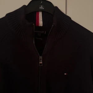 Grå svart kofta från Tommy Hilfiger - Snygg grå svart kofta från Tommy Hilfiger med dragkedja framtill och ribbad hög krage. Diskret logga på bröstet och coolt rött och vitt band i nacken. Fullzip 