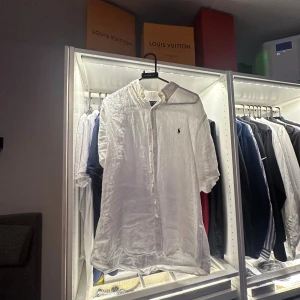 Vit linneskjorta från Polo Ralph Lauren - Fräsch vit skjorta från Polo Ralph Lauren i luftig linne. Custom fit med klassisk krage och knappar framtill. Ikoniska Polo-loggan broderad på bröstet. Perfekt för sommarens alla tillfällen, snyggt avslappnad passform och korta ärmar.