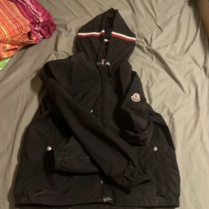 Moncler windbraker - Svart vindjacka från Moncler med huva och dragkedja framtill. Jackan har en vit och röd rand på huvan, Moncler-logga på ärmen och praktiska fickor med tryckknappar. Materialet är lätt och syntetiskt, perfekt för blåsiga dagar.