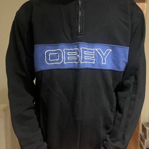 Svart och blå half zip tröja från Obey.  - Svart långärmad tröja från Obey med blå panel och stor vit broderad logga över bröstet. Half zip-dragkedja vid halsen och avslappnad passform. Perfekt för dig som gillar streetwear och vill sticka ut med en snygg detalj.