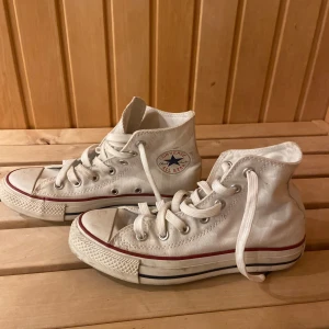 Vita Converse All Star high tops - Klassiska vita Converse All Star high tops med röd och blå rand på sulan. Skorna har snörning, rund tå och är tillverkade i canvas. Perfekta för en avslappnad och cool stil.