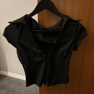 Svart korsett-topp med krage - Snygg svart korsett-topp med korta ärmar och markerad krage. Toppen har djup v-ringning och knäppning framtill med små hakar. Figurnära passform som ger en cool siluett, perfekt för att lyfta vilken outfit som helst.