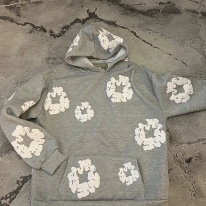 Grå hoodie från Denim Tears med tryck - Grå hoodie från Denim Tears med vita blomliknande tryck över hela tröjan. Tröjan har huva och en klassisk känguruficka framtill. Mjuk insida och avslappnad passform, perfekt för en chill och trendig look.