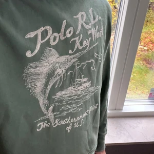 Grön hoodie Polo Ralph Lauren Key West - Snygg grön hoodie från Polo Ralph Lauren med broderad grafik på bröstet som visar en fisk och båt samt texten 'Key West'. Perfekt för dig som gillar chill och sportig stil.