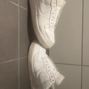 Nike Air Force 1 vita sneakers - Klassiska helvita Nike Air Force 1 sneakers i skinn med perforerade detaljer på tån och den ikoniska swoosh-loggan på sidan. Tidlös modell med platt sula och rund tå, perfekt för dig som gillar en clean och stilren look.