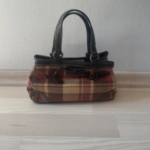 Rutig handväska från Tommy Hilfiger - Jätte snygg vintage handväska från Tommy Hilfiger i rutigt mönster med bruna och beige toner! Väskan har detaljer i mörkbrunt skinn, dubbla handtag och metallringar. Perfekt storlek för det viktigaste och stängs med magnetknapp. Klassisk och tidlös design. Fint att ha i höst