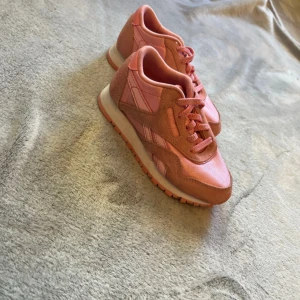 Rosa sneakers från Reebok - Säljer ett par snygga rosa sneakers från Reebok med klassisk design. Skorna har rund tå, platt sula och snörning. Ovandelen är i en mix av mocka och syntetmaterial, vilket ger en cool kontrast. Perfekt för dig som gillar färg och vill sticka ut.