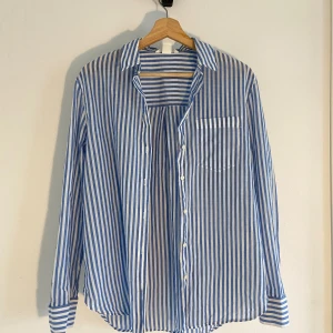Blåvit randig skjorta från H&M - Snygg blåvit skjorta från H&M i storlek 36. Skjortan har vertikala ränder, klassisk krage, bröstficka och knappar framtill. Tillverkad i mjuk bomull, perfekt för en chill och stilren look. Långärmad med manschettknappar och rundad nederkant.