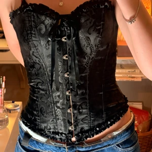 Svart korsett med snörning och mönster - Säljer en svart korsett med snyggt broderat mönster och glansig finish. Den har snörning framtill, dekorativa knappar och volangkant upptill och nedtill. Materialet känns som satin och polyester, perfekt för dig som vill ha en utklätfning till halloween 