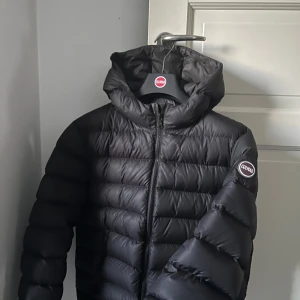 Svart Colmar pufferjacka med huva - Säljer en svart pufferjacka från Colmar med huva och dragkedja framtill. Jackan har ett stilrent, quiltat mönster och Colmar-logga på ärmen. Perfekt för kalla dagar och har en snygg, modern look som funkar till streetwear. Ett lagat hål på armen (se bild) priset kan diskuteras vid snabb affär.  Stl 172. Nypris:4499kr