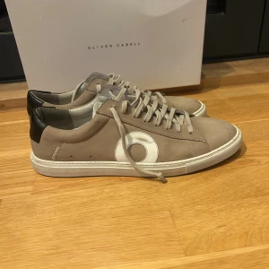 Oliver Cabell skor  - Snygga sneakers från Oliver Cabell i beige mocka med vit rund detalj på sidan och svart häl. Skorna har snörning, platt vit sula och rund tå. Perfekt för dig som gillar stilrena och moderna sneakers med en unik touch. Skick 8,9/10