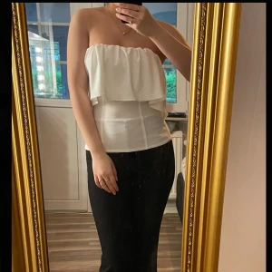 Vit bandeau tubtopp från Zara - Säljer en vit bandeau tubtopp från Zara med volang upptill. Toppen är axelbandslös och har en snygg croppad passform som sitter tight mot kroppen. Helt ny med prislapp!